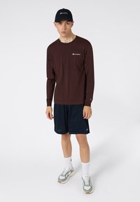 Chemise à manches longues marron avec logo blanc, associée à un short en mesh bleu marine, des chaussettes blanches et des baskets grises avec des accents. La casquette est bleu foncé.