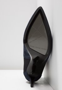 Escarpin à bout pointu en daim bleu marine, talon kitten incliné contre un coin, montrant la semelle noire avec le logo Calvin Klein répété.