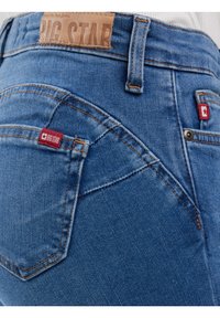 Tuvplāns zilām džinsa biksēm ar redzamu sarkanas Big Star zīmola tagu un ādas zīmola uzliku jostasvietā.