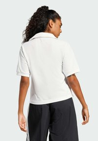 Biela polo košeľa s golierom a prehnutými rukávmi, hladká tkanina, priliehavý strih, zobrazená zozadu, v kombinácii s voľnými čiernymi nohavicami.