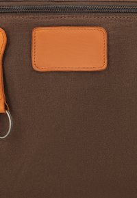 Bruin stoffen oppervlak met een oranje leren patch en een gedeeltelijk zichtbare oranje leren sleutelhanger met metalen ring nabij de rits.