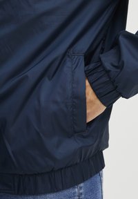 Marineblauwe jas van lichtgewicht, glad materiaal; voorzien van elastische mouwen en een zijzak. Inclusief gathering aan de onderkant.
