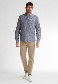 Camicia button-up a scacchi blu chiaro e bianco, con maniche lunghe e tasca sul petto, abbinata a pantaloni khaki e sneakers bianche.
