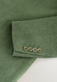 Groene getextureerde blazer mouw met vier beige knopen op de manchet, getoond op een witte ondergrond.