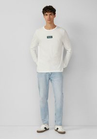 White long-sleeve shirt with a green logo on the chest, paired with light blue jeans and white sneakers with a dark accent.