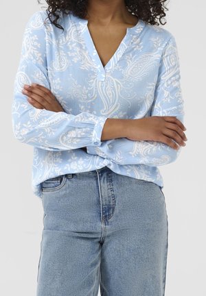 Kobieta ze skrzyżowanymi ramionami, ubrana w jasnoniebieską bluzkę w paisley oraz jasnoniebieskie dżinsy, na jednolitym tle.
