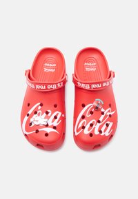 Κόκκινα Crocs με την επωνυμία Coca-Cola και λευκό λογότυπο, ρυθμιζόμενο λουράκι με τη φράση «It’s the real thing» και δύο μικρά διακοσμητικά καρφίτσες στην κορυφή.