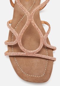 Sandalia en oro rosa con adornos, tiras entrelazadas, plantilla de ante y suela plana. Presenta detalles brillantes y una textura suave.