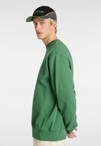 Sudadera verde de corte relajado, con cuello redondo y puños de canalé. Combinada con una gorra negra y gris con detalles en verde y amarillo.