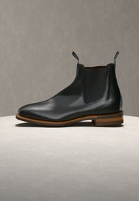 Botte Chelsea en cuir noir poli avec panneaux latéraux élastiques, languettes de traction et talon empilé en bois sur une surface en pierre claire avec un fond gris uni.