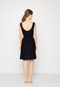 Vestido preto sem mangas com costas baixas, cintura ajustada e bainha ampla. Feito de um tecido macio, combinado com sandálias de salto de cor clara.