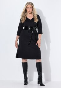 Abito nero fino al ginocchio con un motivo texturizzato, scollo a V, maniche a tre quarti e una cintura nera lucida. La modella indossa stivali neri alti.