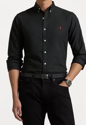 Uomo che indossa una camicia nera a maniche lunghe con logo rosso ricamato, cintura nera, pantaloni neri e orologio da polso con cinturino nero.