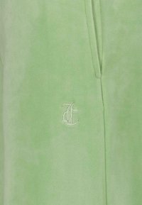 Vêtement en velours vert clair avec une texture lisse. Présente un petit logo brodé. Le design comprend des détails de couture sur les côtés.