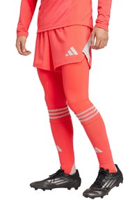 Heldroze sportoutfit bestaande uit een langemouwen top en shorts, gecombineerd met roze leggings met drie witte strepen en zwarte voetbalschoenen.