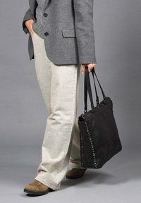 Zwarte lederen tote bag met studs aan één kant, vastgehouden door zwarte hengsels. Gecombineerd met een grijze blazer en lichtbeige broek. Amandelvormige bruine schoenen.