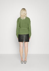 Grüner gestrickter Pullover mit weiten Ärmeln und einem Taillengürtel, kombiniert mit einem schwarzen Leder-Minirock und hochhackigen, gemusterten Sandalen.