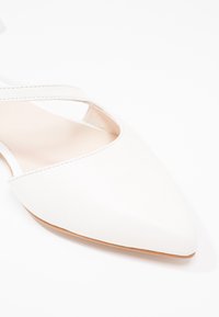Chaussure plate blanche à bout pointu avec une bride étroite sur le dessus et une semelle marron clair, présentée sur fond blanc.