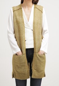 Olivgrön, lång stickad väst med en mjuk textur, öppen fram och med två framfickor. Bärs över en vit topp och mörka jeans.