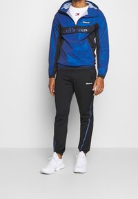 Blue and black windbreaker with a half-zip and logo, paired with black joggers featuring blue side stripes; worn with white sneakers.