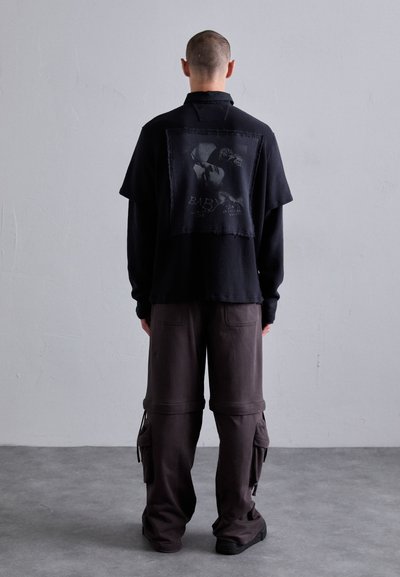 Man with shaved head stands facing gray wall wearing black long-sleeve top with graphic back patch and dark cargo pants with pockets.