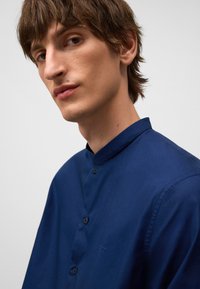 Donkerblauwe shirt met een staande kraag, voorzien van een knoopsluiting en een gladde textuur. Minimalistisch ontwerp met contrasterende zwarte knopen.