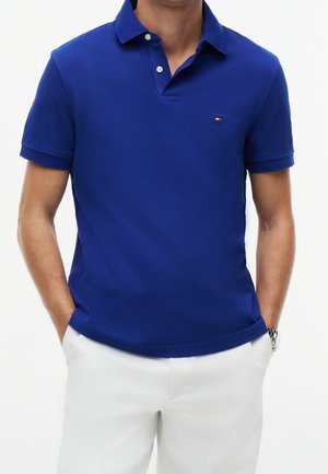 Hombre que lleva una camiseta polo de manga corta de color azul real con dos botones y pantalones blancos, manos en los bolsillos, reloj de pulsera en la muñeca izquierda.