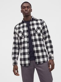 Homme portant une chemise à carreaux noir et blanc boutonnée sur un t-shirt noir avec un pantalon gris, debout devant un fond clair uni.