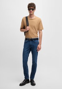 Tan short-sleeve t-shirt, dark blue jeans, black belt, and black shoes, with a leather bag and sunglasses. Smooth texture, fitted design.