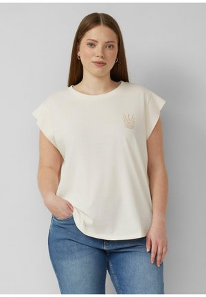 Frau mit glattem braunem Haar, die ein weißes ärmelloses Oberteil mit kleinem Sonnenmotiv und blaue Jeans trägt, steht mit einer Hand in der Tasche.