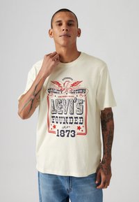 Beige katoenen t-shirt met een grafisch ontwerp met rode en marineblauwe tekst: "LEVI'S FOUNDED 1873" en een adelaarontwerp. Korte mouwen, ontspannen pasvorm.