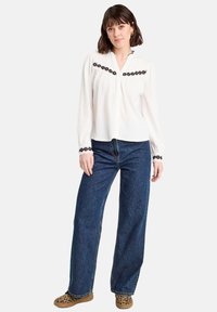 Blouse blanche à manches longues avec un tissu texturé et une broderie florale noire sur la poitrine et les poignets, associée à un jean bleu à jambes larges et des chaussures à imprimé léopard.