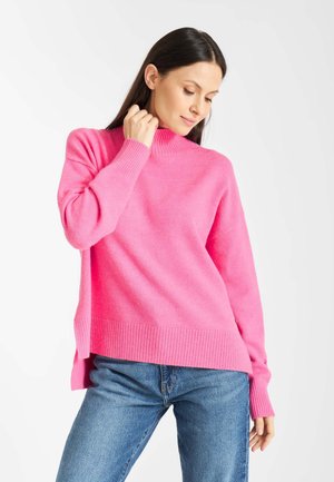 Roze gebreide trui met een hoge hals, geribbelde manchetten en zoom, en een ontspannen pasvorm. Gecombineerd met blauwe jeans, wat een casual en comfortabele stijl laat zien.