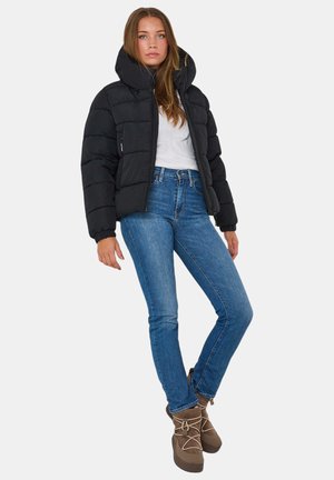 Schwarze Daunenjacke mit hohem Kragen, Reißverschluss und strukturiertem Design, kombiniert mit blauen Jeans und braunen Schnürstiefeln.