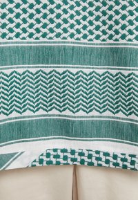 Un tissu à motifs verts et blancs présente des motifs géométriques, notamment des losanges et des zigzags, avec un tissage texturé et des rayures horizontales.
