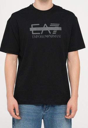 Mann trägt ein schwarzes T-Shirt mit kurzen Ärmeln, großem grauem "EA7 Emporio Armani"-Logo und blauer Jeans, steht vor einem einfarbigen Hintergrund.