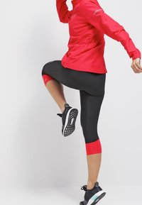 Persona con chaqueta roja y leggins negros con acentos rojos levanta una pierna, mostrando zapatillas de running en blanco y negro contra un fondo blanco.