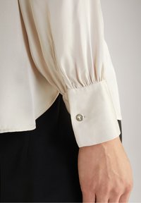 Blusa color beige chiaro con design a maniche arricciate e polsino con bottoni, dotata di un bottone d'accento argentato e texture di tessuto liscia.