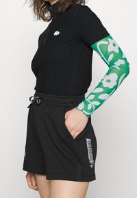 Black ribbed short-sleeve top with a zipper, featuring a white logo. Paired with black shorts, accented by a green floral-patterned sheer sleeve.