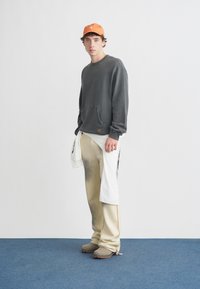 Felpa grigia con tasca frontale; pantaloni crema e beige con motivo sbiadito; berretto arancione; scarpe taupe con suola in gomma.