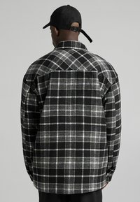 Svart och grå rutig flanellskjorta med klassisk skärning, detaljerad sömnad och en spetsig krage; bärs med en svart keps bak och fram.