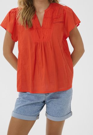 Kobieta ubrana w jasnopomarańczową, krótki rękaw, plisowaną bluzkę oraz jasnoniebieskie, podwinięte szorty dżinsowe, stojąca na białym tle.