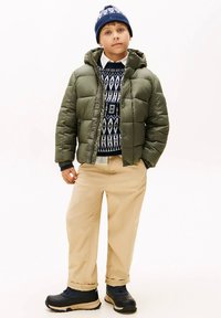 Giacca a pelo verde con cappuccio, maglione a fantasia blu navy e bianco sotto, pantaloni beige e stivali neri con suola color cuoio. Cappello di lana blu in testa.