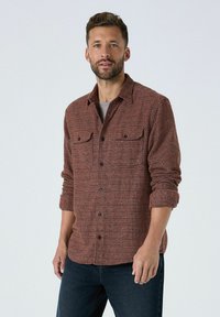 Lühi juustega ja habemega mees, kes kannab tekstureeritud pruuni nööbiga särki, mille varrukad on üles keeratud, ning tumedaid siniseid teksaseid lihtsa tausta ees.