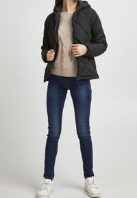 Svart vadderad pufferjacka med huva, beige stickad tröja, mörka skinny jeans och vita sneakers, stående mot en enkel bakgrund.