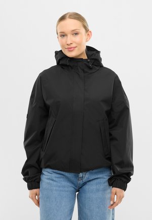 Femeie îmbrăcată într-o geacă neagră cu glugă, buzunare frontale și manșete elastice, purtând blugi bleu deschis, stând pe un fundal simplu.