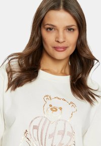 Crème-kleurige sweatshirt met een berenprint in roségoud, voorzien van hartvormige details en een gestructureerd ontwerp.