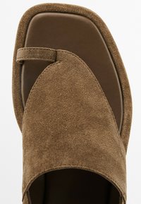 Sandale en daim marron avec une large bride, un bout carré et un fin détail en tanga. Possède un intérieur lisse de couleur marron foncé. Design simple et plat.