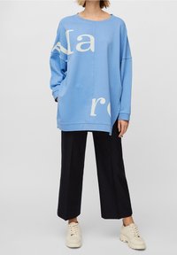 Blå oversized sweatshirt med vit grafisk text, kombinerad med vida svarta byxor och ljusbeiga skor. Mjuk tyg, avslappnad passform, rund hals.