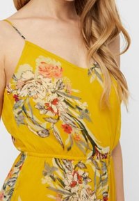 Robe jaune à fleurs avec un col en V, des bretelles fines et une taille élastique. Le tissu est léger avec une texture douce et des motifs floraux multicolores.